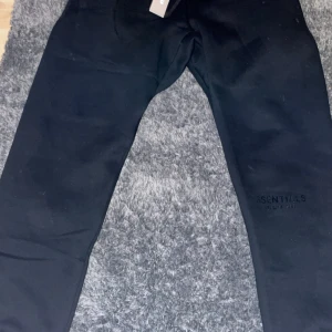 Svarta Essentials Fear of God mjukisbyxor - Svarta mjukisbyxor från Essentials Fear of God med elastisk midja och snörning. Byxorna har en bakficka och diskret logga på benet. Sköna och stilrena, perfekta för chill eller streetwear. Materialet är mjukt och känns riktigt nice mot huden.