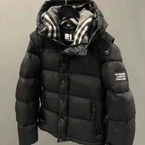 Svart pufferjacka från Burberry - Säljer en svart pufferjacka från Burberry med klassisk logotyp på ärmen och rutigt foder i huvan. Jackan har dragkedja och knappar framtill, samt praktisk huva med snörning. Perfekt för kalla dagar och riktigt snygg streetstil.