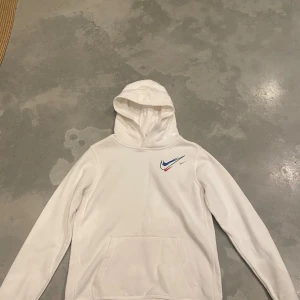 Vit hoodie från Nike med färgglada loggor - Säljer en clean vit hoodie från Nike med flera färgglada swoosh-loggor på bröstet i blått, grönt, rött och silver. I STORLEK 158-170 CM. Tröjan har huva och en stor magficka. Perfekt för dig som gillar sportig och stilren streetwear.