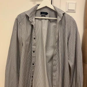 Randig skjorta från Riley, regular fit - Snygg skjorta från Riley i regular fit med smala svarta och vita ränder. Klassisk krage, långa ärmar och knäppning framtill. Perfekt för dig som gillar stilrena och tidlösa plagg.