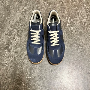 MM Gats - Snygga sneakers från Maison Margiela i mörkblå mocka och skinn med vita snören och beige gummisula. Klassisk retrostil med rund tå och kontrasterande material. Perfekta för dig som gillar stilrena och trendiga skor med en twist.