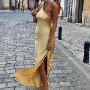 Gul långklänning med slits och öppen rygg - Säljer en gul långklänning i satinliknande material med oneshoulder och smala axelband. Klänningen har en snygg slits framtill och en öppen rygg med dekorativt band. Passformen är slim och tyget har en fin glans. Perfekt för sommarkvällar och speciella tillfällen.