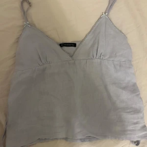 Ljusgrå linnetopp från Brandy Melville - Supersöt ljusgblå linnetopp från Brandy Melville med smala justerbara axelband och v-ringning. Toppen har små rosettdetaljer vid axelbanden och ett knytband i ryggen för extra passform. Deras onesize storlek så skulle säga att denna är som en xs/s💕💕🥰