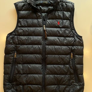 Svart dunväst från Polo Ralph Lauren - Snygg svart dunväst från Polo Ralph Lauren med quiltad design och dragkedja framtill. Västen har två fickor med dragkedja och klassisk röd logga broderad på bröstet. Perfekt att slänga över en hoodie eller tröja för en chill look. Tvär fet väst från Ralph lauren köpt i USA! Hör av dig vid minsta fundering MVH Leo