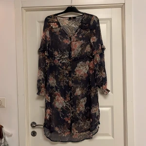 Blommig klänning/tunnika från Capri Collection - Säljer en transparent, blommig klänning/tunika från Capri Collection i marinblått med inslag av rosa, beige och vitt. Klänningen har v-ringning, långa ärmar med volangdetaljer och ett luftigt, flowy material. 