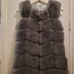 Mörkgrå fluffig pälsväst - Säljer en riktigt snygg mörkgrå pälsväst med fluffig och mjuk känsla. Västen har en rak och lite oversized passform, med breda paneler som ger en cool och lyxig look. Perfekt att slänga över en hoodie eller långärmad tröja för extra värme och stil. Defekt i bak pga ett tuggummi som fastnat och avlägsnats.i övrigt bästa skick. 