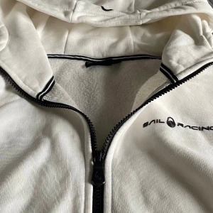 Vit hoodie från Sail Racing med dragkedja - Snygg vit hoodie från Sail Racing med svart dragkedja och broderad logga på bröstet. Tröjan har huva, långa ärmar och ribbade detaljer med svarta och vita ränder vid halsen. Mjuk insida i fleece som gör den extra skön. Perfekt för chill dagar. Kom dm för frågor!