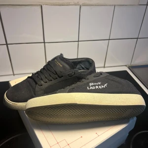 Svarta Saint Laurent sneakers - Svarta sneakers från Saint Laurent med vit logga på sidan och guldfärgad text på plösen. Skorna har snörning, rund tå och en vit platt sula. Tillverkade i canvas med detaljer i skinn. Perfekt för dig som gillar stilrena och exklusiva sneakers.