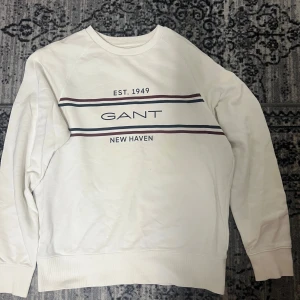 Vit sweatshirt från GANT XL - Vit sweatshirt från GANT i storlek XL med tryck 'EST. 1949 GANT NEW HAVEN' på bröstet. Tröjan har långärmad design, ribbade muddar och klassiska ränder i marinblått och vinrött över bröstet. Tillverkad i mjuk bomull för skön komfort.