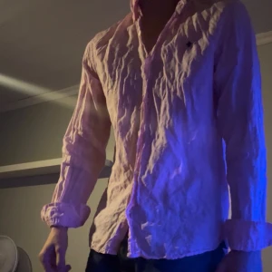 Ljusrosa skjorta med broderad logga - Säljer en ljusrosa skjorta från Morris med klassisk krage och knappar framtill. Skjortan har lång ärm och en snygg broderad logga på bröstet. Tillverkad i mjuk bomull som känns skön mot huden. Perfekt för dig som gillar stilrena plagg med en twist.