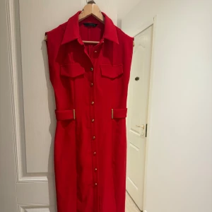 Zara klänning - Snygg röd ärmlös klänning från Zara med markerad krage, guldknappar framtill. Klänningen har midjedetaljer med guldfärgade spännen och en slits fram (se sista bild). Använt 1 gång