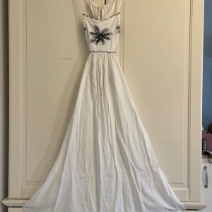 Vit långklänning med broderi - Vit långklänning med svart broderad blomdetalj på bröstet och axelbandslös design. Klänningen har en markerad midja och ett luftigt, svepande fall. Perfekt för sommarens alla tillfällen.