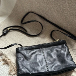 Svart axelväska i skinn från Wera - Snygg svart axelväska från Wera i mjukt skinn med guldfärgade dragkedjor och detaljer. Väskan har ett stilrent och minimalistiskt utseende, smidig storlek och justerbar axelrem. Perfekt för dig som gillar klassisk design med en touch av lyx.