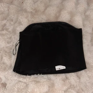 Svart tubtopp med dragkedja - Säljer en stilren svart tubtopp med rak passform och diskret dragkedja i sidan för enkel på- och avtagning. Toppen är axelbandslös och har en slät yta, perfekt för att skapa en clean look. Materialet känns mjukt och följsamt mot huden.