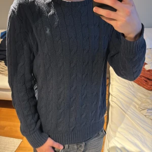 Mörkblå kabelstickad tröja Polo Ralph Lauren - Säljer en mörkblå kabelstickad tröja från Polo Ralph Lauren med klassisk rund halsringning och diskret broderad logga på bröstet. Tröjan har ribbade muddar vid ärmslut och nederkant, perfekt för en stilren och avslappnad look.