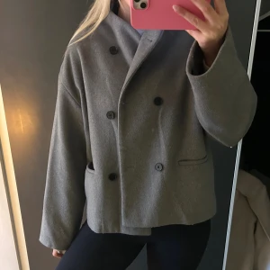 Kort kappa från NA-KD - Säljer en snygg grå jacka från NA-KD i storlek 36, passar S/M. Jackan har dubbelknäppning med svarta knappar, klassisk krage och två fickor framtill. Sällan använd så är i superfint skick. 