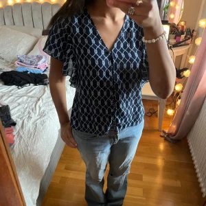 Mörkblå blus med mönster från Gina Tricot - Säljer en mörkblå blus från Gina Tricot i storlek 36. Blusen har ett vitt rutigt mönster med knutdetaljer, v-ringad hals och knappar framtill. Ärmarna är korta och har en lätt volang.