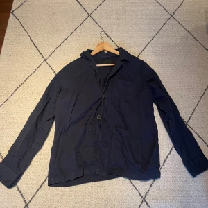 Mörkblå overshirt från 157 - Säljer en mörkblå overshirt från 157 med klassisk krage, två stora fickor framtill och en bröstficka. Jackan har knappar och är långärmad, perfekt för lager-på-lager. 