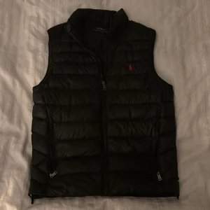 Svart dunväst från Polo Ralph Lauren XS - Svart dunväst från Polo Ralph Lauren i storlek XS. Västen har quiltad design, dragkedja framtill och två sidofickor med dragkedja. Klassisk röd Polo-logga broderad på bröstet. Perfekt att slänga över en hoodie för en chill look.