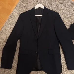 Mörkblå kostym från Zara, storlek 46 - Snygg mörkblå kostym från Zara i klassisk modell med kavaj och matchande kostymbyxor. Kavajen har två framfickor med lock, smal krage och knäppning med en knapp. Byxorna är raka och har bälteshällor. Perfekt för dig som gillar stilrena och enkla plagg.