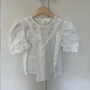 Vit blus med broderad spets Gina Tricot - Säljer en vit blus från Gina Tricot med puffiga korta ärmar och rund hals. Framtill har den en snygg broderad spetsdetalj i triangelform som ger en romantisk vibe. Blusen är gjord i mjuk bomull och har knappstängning bak i nacken.