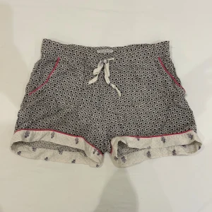 Mönstrade shorts från Scotch & Soda - Snygga pyjamas shorts från Scotch & Soda med svart och vit geometriskt mönster, kontrasterande rosa kant och snörning i midjan. Uppvikta ben med ljusare tyg och små tryckta motiv. 