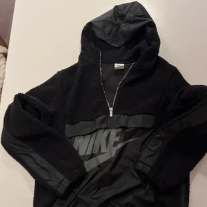 Svart teddyjacka med huva från Nike - Svart teddyjacka från Nike i storlek XL, med stor logga framtill och mindre logga på ryggen. Jackan har huva, half zip och detaljer i syntetmaterial på ärmar och bröst. Perfekt för dig som gillar sportig och chill stil.
