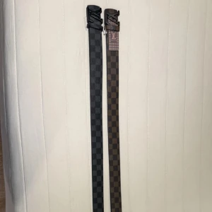 Louis Vuitton rutiga bälten - Två snygga bälten från Louis Vuitton med klassiskt rutigt mönster. Ett i mörkgrått och svart, det andra i brunt och beige. Båda har LV-logga på spännet och är tillverkade i skinn. Perfekt accessoar för att lyfta din outfit.