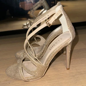 Glittriga guldiga sandaler med klack - Säljer ett par glittriga sandaler från Tiamo i guld med höga klackar och smala remmar som korsar över foten. Skorna har öppen tå och spänne vid ankeln. Perfekta för att sticka ut med en extra touch av glamour. Använda endast en gång. 