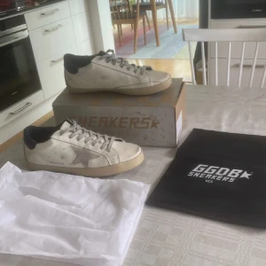 Golden Goose Superstar sneakers - Golden Goose Superstar sneakers i vit med grå stjärndetalj på sidan och mörkblå häl. Skorna har snörning, platt sula i beige och rund tå. Materialet är skinn med mockadetalj på stjärnan. Snyggt slitna och levereras med originalkartong och dustbag. Knappt använda 