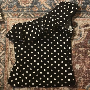 Svart prickig oneshoulder topp med volang - Säljer en svart oneshoulder topp med vita prickar och en stor volang längs axeln. Toppen har en tight passform och är tillverkad i ett mjukt syntetmaterial. Perfekt för dig som gillar att sticka ut med coola detaljer och mönster.