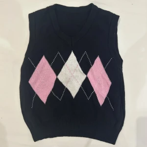 Svart rutig stickad väst i bomull - Svart stickad väst i 100% bomull med klassiskt rutig argyle-mönster i rosa och vitt på framsidan. V-ringad hals och ribbad nederkant ger en snygg och chill look. Perfekt att styla över en skjorta eller t-shirt för en preppy vibe.