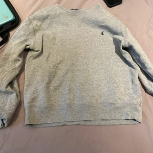Grå sweatshirt från Polo Ralph Lauren - Klassisk grå sweatshirt från Polo Ralph Lauren med rund halsringning och broderad marinblå logga på bröstet. Tröjan har långa ärmar och är tillverkad i mjuk bomullsmix, perfekt för en chill och stilren look.