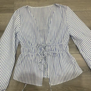 Blårandig blus med snörning - Säljer en blårandig blus med v-ringning och snörning i midjan. Blusen har långa ärmar och ett luftigt, loose fit. Perfekt för dig som gillar en chill och stilren look med lite extra detaljer.