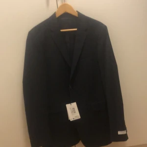 Mörkblå kavaj från Tiger of Sweden - Snygg mörkblå kavaj från Tiger of Sweden med klassisk passform och stilrena knappar. Kavajen har märkets logga på insidan och vid ärmslutet. Perfekt för dig som vill ha en stilren och tidlös look. Kavajen är helt ny med prislappen kvar 