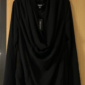 Svart omlottklänning från Boohoo - Svart kortklänning från Boohoo med omlott-drapering framtill och transparent meshdetalj vid nederkanten. Klänningen har lång ärm och en snygg, flödande siluett som ger en cool vibe. Perfekt för dig som gillar stilrena och trendiga plagg.