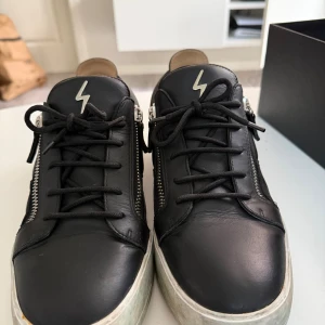 Svarta sneakers Giuseppe Zanotti - Svarta sneakers från Giuseppe Zanotti med cool blixtdetalj på plösen och silverfärgade dragkedjor på sidorna. Skorna har snörning, rund tå och vit platt sula med slitningar. Tillverkade i skinn med beige insida. Snygg och edgy design.