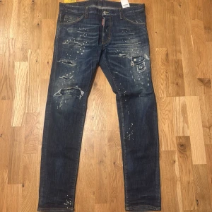 Dsquared2 Cool Guy jeans blå, stl 50 - Snygga Dsquared2 Cool Guy jeans i mörkblå tvätt med slitningar, färgstänk och trasiga detaljer. Klassisk femficksmodell med raka ben och orange kontrastsömmar. Märkespatch och etikett fram. Perfekt för dig som gillar street och edgy stil.