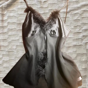 Svart satin topp med fjädrar - Svart topp i satin med tunna axelband och dekorativa fjädrar längs urringningen och framtill. Klänningen har en lätt glansig finish och en feminin siluett som ger en lyxig känsla. Perfekt för dig som gillar att sticka ut med detaljer.