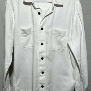 Vit overshirt med svarta knappar - Vit overshirt med klassisk krage och två stora bröstfickor. Skjortan har svarta knappar längs hela framsidan och är tillverkad i ett mjukt bomullstyg. Långärmad och med en loose passform.