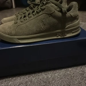 Olivgröna sneakers från Polo Ralph Lauren storlek 36 - Snygga olivgröna sneakers från Polo Ralph Lauren med guldtext på hälen. Skorna är tillverkade i mocka med snörning och har en platt sula. Diskret Polo-logga på sidan och Ralph Lauren-tryck på sulan. Perfekt för dig som gillar stilrena och klassiska sneakers.