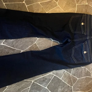 Mörkblå bootcut jeans med fickdetaljer - Snygga mörkblå jeans med bootcut passform och klassiska fickor med lock och knapp bak. Jeansen har kontrastsömmar och är tillverkade i ett robust denimtyg. Perfekta för dig som gillar en tidlös och clean look. Jag har använt den max 1 gång.