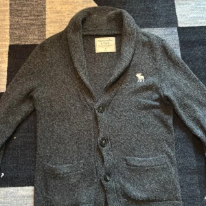 Mörkgrå kofta Abercrombie & Fitch - Mysig mörkgrå kofta från Abercrombie & Fitch med två framfickor och klassisk sjalkrage. Tröjan har långa ärmar, knäppning med tre knappar och en vit broderad älg på bröstet. Perfekt för lager-på-lager och chill stil. Aldrig använd 