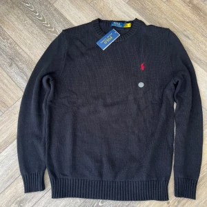 Svart stickad tröja Polo Ralph Lauren - Svart stickad tröja från Polo Ralph Lauren med klassisk rund hals och ribbade muddar. Tröjan har ett rött broderat Polo-logo på bröstet och är långärmad. Perfekt för dig som gillar stilrena och tidlösa plagg.