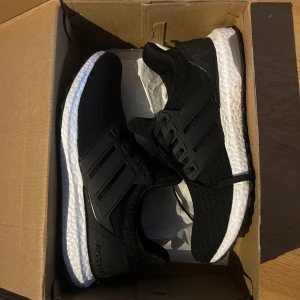 Adidas Ultraboost svarta sneakers - Svarta Adidas Ultraboost sneakers med stickad ovandel och klassiska tre ränder på sidorna. Skorna har en vit, responsiv Boost-sula och svarta detaljer i syntet. Perfekta för dig som vill ha både stil och komfort i vardagen.