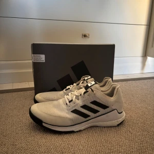 Adidas Crazyflight vita sneakers - Adidas Crazyflight sneakers i vit med svarta detaljer och klassiska tre ränder på sidan. Skorna har snörning, platt sula och en lätt, ventilerande ovandel i syntetmaterial. Perfekta för inomhussport och träning, med bekväm Boost-dämpning.