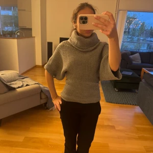 Grå stickad polotröja med tajt midja - Säljer en grå stickad polotröja med bred och hög krage. Tröjan har korta, vida ärmar och en ribbad, tight midja som ger en snygg siluett. Perfekt för dig som gillar chill och stilren look. Passar till svarta jeans eller kostymbyxor.