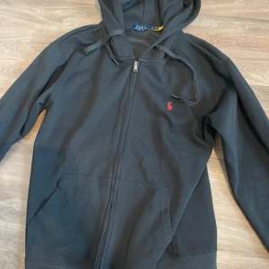 Svart hoodie från Polo Ralph Lauren - Svart hoodie från Polo Ralph Lauren med röd logga på bröstet. Tröjan har huva med snörning, dragkedja framtill och två fickor. Tillverkad i mjuk bomull, perfekt för chill dagar. Klassisk och stilren design som funkar till det mesta.