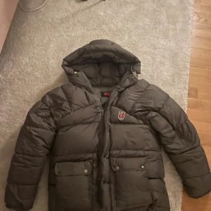 Mörkgrön  pufferjacka från Fjällräven - Säljer en mörkgrön pufferjacka från Fjällräven i storlek XXXS i unisex men passar som S-xxs Jackan har huva med snörning, två stora fickor med knappar och dragkedja framtill. Perfekt för kalla dagar och har den klassiska Fjällräven-loggan på bröstet. På sista bilden är ett jagat hål