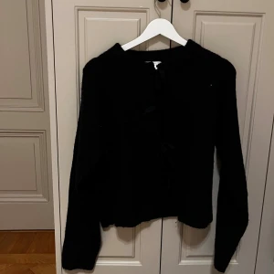 Svart kofta med rosetter från Gina Tricot - Mysig svart kofta från Gina Tricot med lång ärm och ribbad krage. Framsidan har tre snygga rosetter som ger en unik touch. Koftan är stickad och har en loose passform, perfekt för att hålla sig varm och ändå se cool ut.